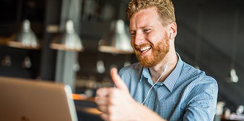  Ein lächelnder, bärtiger Mann mit Ohrhörern und kariertem Hemd gibt einen Daumen hoch. Vor ihm ein Laptop in einem modernen Ambiente.