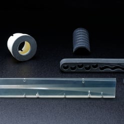  Das Bild zeigt vier verschiedene industrielle Kunststoff- oder Gummiteile auf dunklem Hintergrund: eine Rolle, ein geripptes Halbrund, eine gelochte Leiste und ein transparentes Profil.