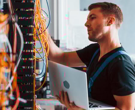 Ein IT-Techniker in Arbeitskleidung hält einen Laptop in der Hand und überprüft konzentriert die Verkabelung in einem Serverschrank mit zahlreichen gelben Netzwerkkabeln.