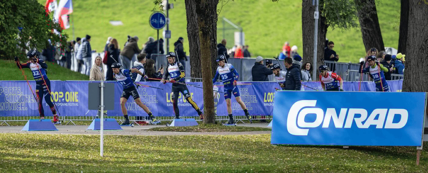 Mehrere Biathleten auf Rollskiern fahren während eines Wettkampfs auf einer asphaltierten Strecke an Zuschauern und blauen Werbebannern in einem Park vorbei.