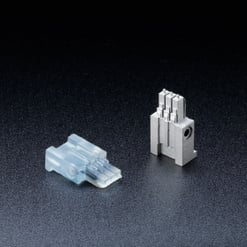  Das Bild zeigt zwei kleine, präzise gefertigte elektronische Bauteile auf dunklem, strukturiertem Hintergrund. Eines ist transparent-weiß, das andere metallisch grau.