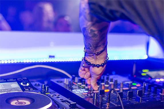 Szene in einem Club. Zu sehen sind ein DJ Controller und eine Hand, die Regler betätigt. Im Hintergrund sind Silhouetten von Menschen zu sehen, die vor dem DJ-Pult stehen. Die Szene ist in blauviolettes Discolicht getaucht.