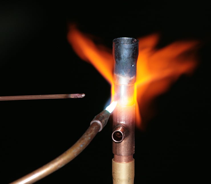Eine Lötflamme erhitzt ein Kupferrohr, während ein Lötdraht an die erhitzte Stelle geführt wird.
