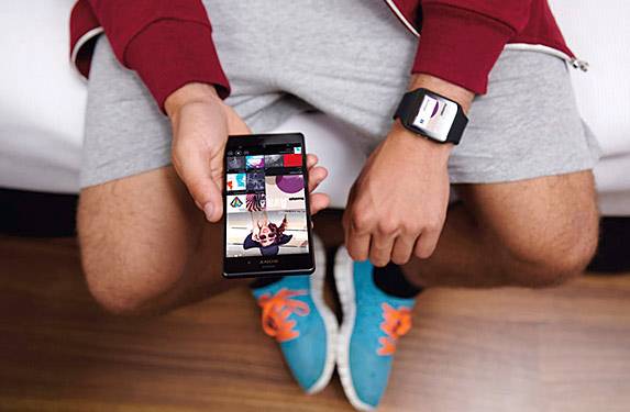 Ein Mann in Sportklamotten sitzt auf einem Sofa oder Bett. Sein Gesicht ist nicht zu sehen. Die Kamera ist auf die Hände fokussiert. Am linken Handgelenk trägt der Mann eine Smartwatch, in der rechten Hand hält er ein Smartphone.