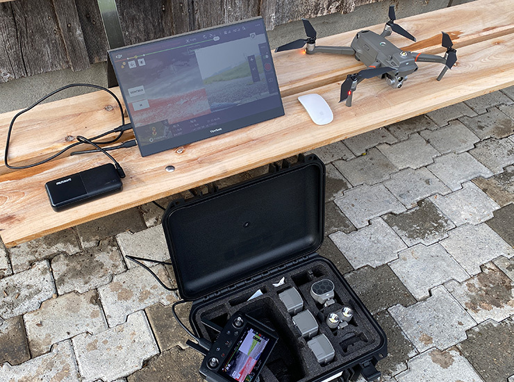 Mobiles Drohnen-Set mit Monitor, Fernbedienung, Akkus und Drohne auf einer Holzbank im Außenbereich.
