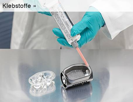  Eine Person in weißem Laborkittel und türkisfarbenen Handschuhen trägt einen hellbraunen Klebstoff aus einer Tube auf ein silbernes Metallobjekt auf, während daneben ein transparentes, hantelförmiges Plastikteil liegt; der Text "Klebstoffe »" im oberen linken Bereich des Bildes weist auf eine Weiterleitung hin.