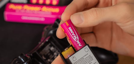 Nahaufnahme einer Hand, die eine pinkfarbene Ansmann-Batterie in das Batteriefach eines schwarzen Geräts einsetzt.