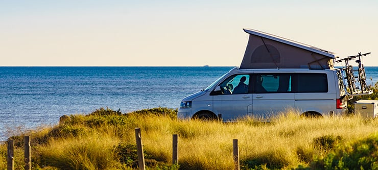 Ein Campingbus mit ausgeklapptem Dach steht am Meer, im Hintergrund ist der Horizont sichtbar.