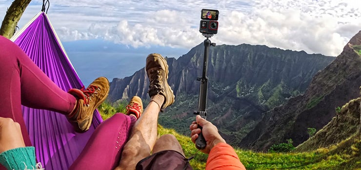 Zwei Personen entspannen in einer Hängematte mit Blick auf eine Berglandschaft, während eine Actioncam auf einem Selfie-Stick gehalten wird.