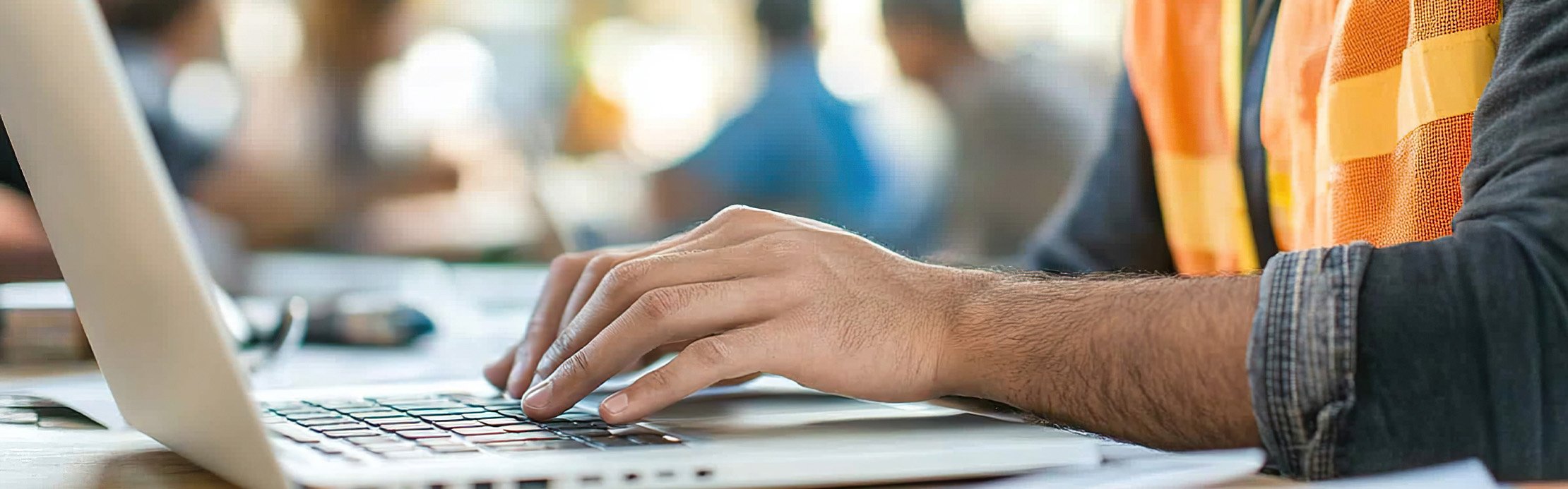 Ein Arbeiter mit einer Leuchtweste sitzend an einem Tisch mit einem Laptop.