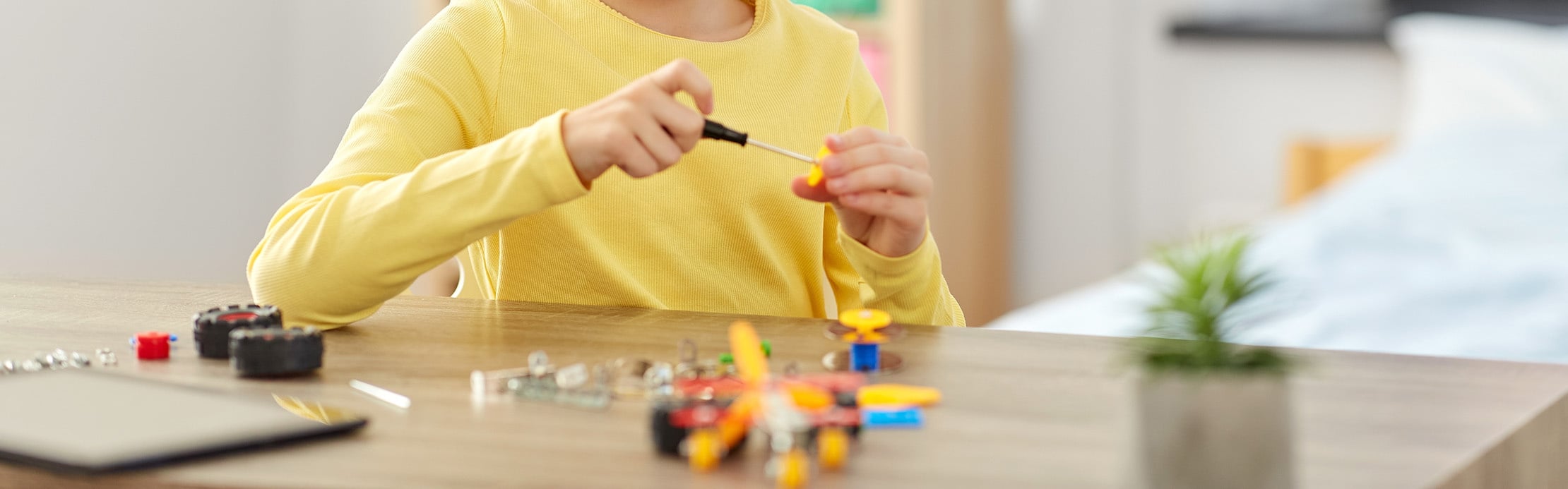Ein Kind im gelben Pullover montiert mit einem Schraubenzieher konzentriert ein buntes Konstruktionsspielzeug an einem Holztisch.