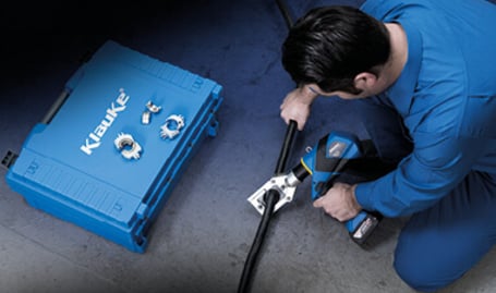 Ein Elektriker kniet neben einem offenen blauen Werkzeugkoffer und bedient ein blaues Akku-Universalwerkzeug an einem Kabel auf dem Boden.