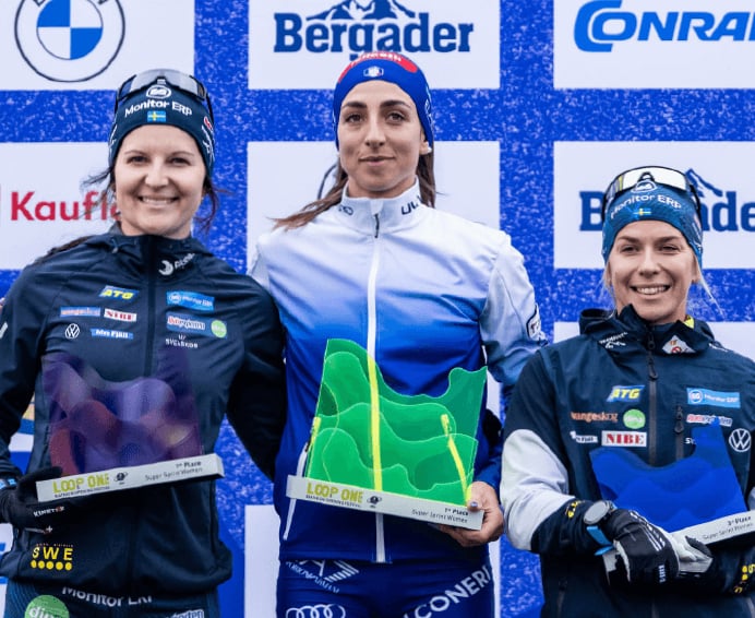 Drei lächelnde Biathletinnen stehen bei einer Siegerehrung vor einer Sponsorenwand und präsentieren ihre gläsernen Trophäen.