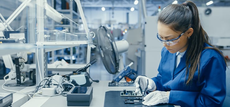 Frau in blauer Arbeitskleidung und weissen Handschuhen arbeitet an einer Elektronikplatine in einer modernen Fabrikumgebung