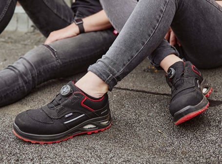 Zwei Personen in grauen Jeans sitzen auf dem Boden, wobei im Vordergrund schwarze ELTEN-Sicherheitsschuhe mit roten Akzenten und BOA-Drehverschluss detailreich präsentiert werden.