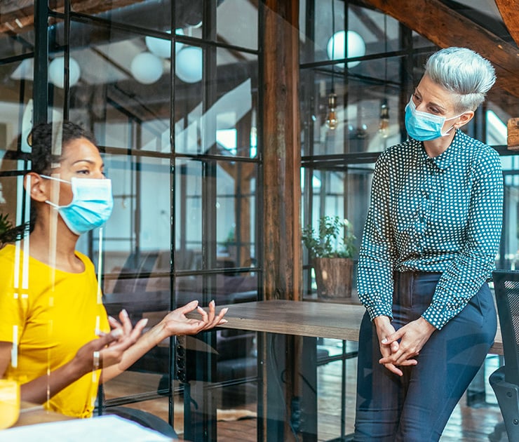 Zwei Frauen mit Gesichtsmasken führen ein Gespräch in einem Büro, getrennt durch eine transparente Schutzscheibe.