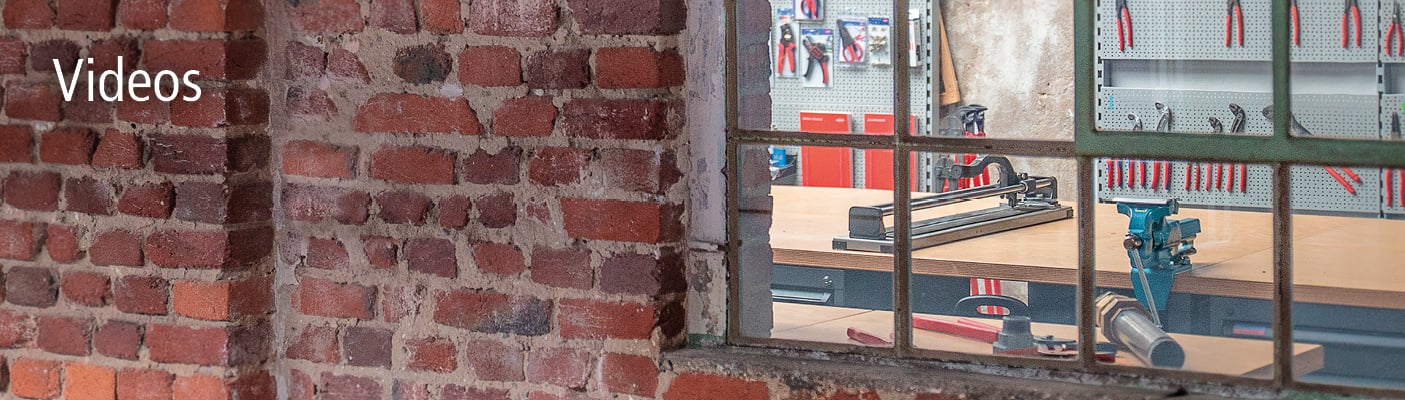 Ein Blick durch ein altes Fenster mit Metallrahmen in eine Werkstatt mit Werkzeugen und einem Fliesenschneider. Auf der Ziegelwand steht 'Videos'.