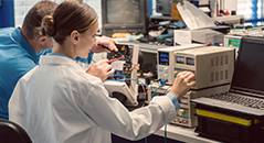 Zwei Personen in einer Laborumgebung untersuchen und reparieren sorgfältig elektronische Geräte, wobei die Person im Vordergrund einen weißen Laborkittel trägt und rote Prüfspitzen an ein Bauteil hält.