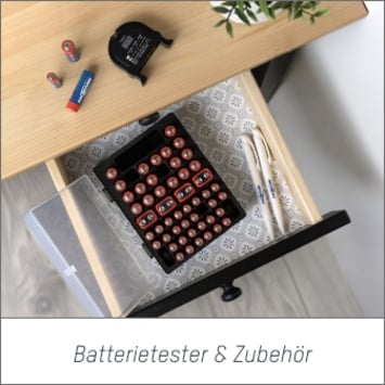 Das Bild zeigt einen Schreibtisch in der Draufsicht mit einer geöffneten Schublade. Auf der Tischplatte liegt ein Batterietester und verschiedene Batterien. Bei Klick auf das Bild werden die Ansmann Batterietester und Zubehör aufgerufen.