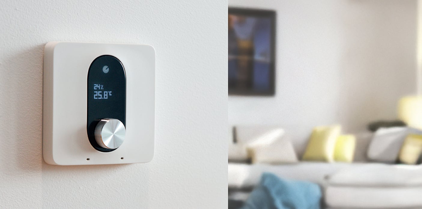 Das Bild zeigt den Shelly LinkedGo smartes Heizungsthermostat an einer Wand montiert. Im Hintergrund ist schemenhaft ein Wohnzimmer zu sehen.