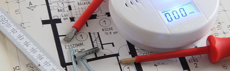 Ein Grundrissplan auf einem Tisch mit verschiedenen Schraubenziehern, einem Massstab und einer Brandmeldeanlage. Ebenfalls befinden sich zwei Schrauben und ein Dübel auf dem Tisch.