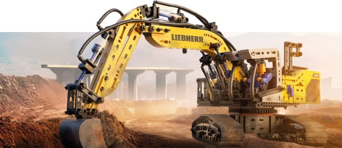 Ein großer, gelber fischertechnik Bagger auf einer Baustelle. Im Hintergrund sind Brückenpfeiler und eine sonnige, staubige Landschaft zu sehen.