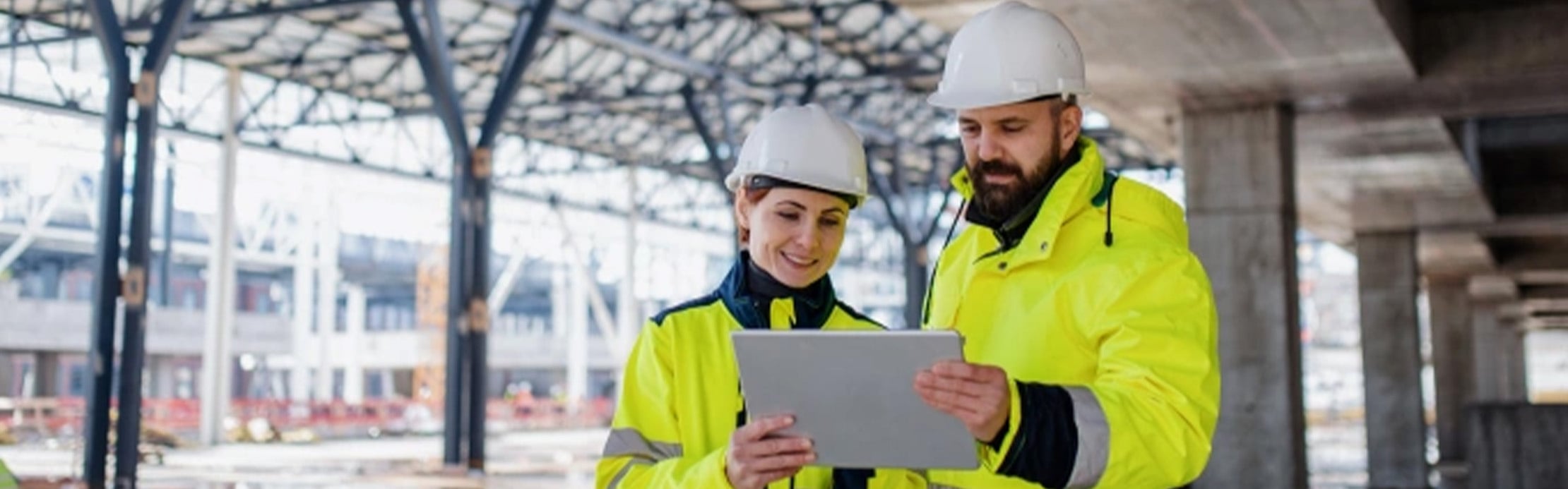 Zwei Fachkräfte mit Warnkleidung und Schutzhelmen prüfen gemeinsam Informationen auf einem Tablet auf der Baustelle