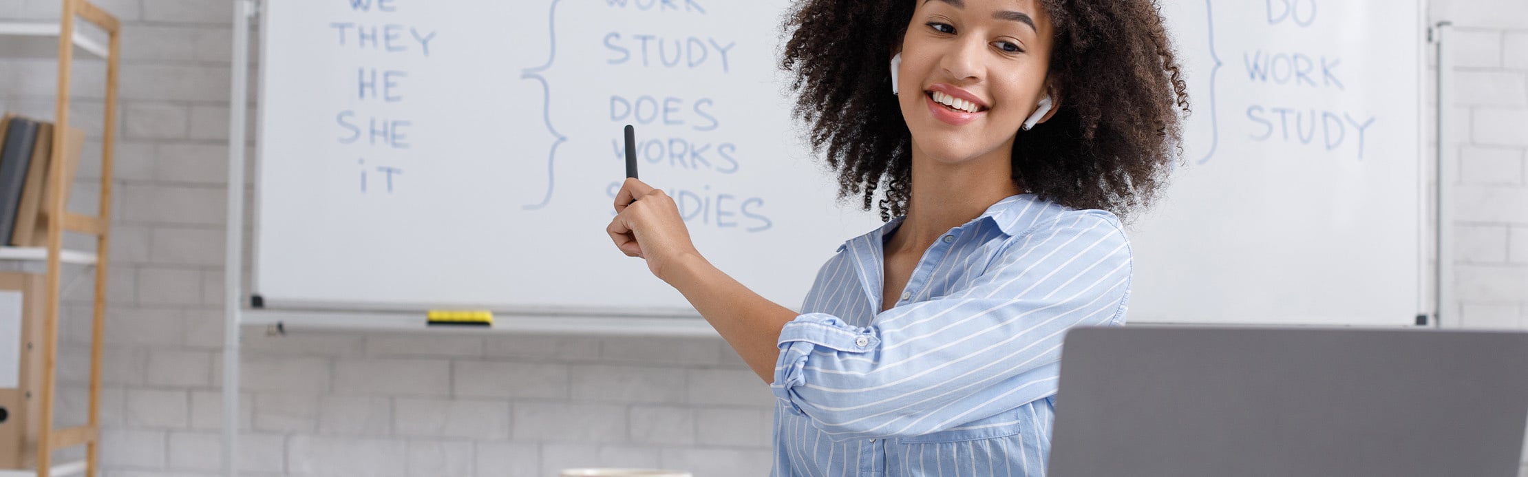 Junge Lehrerin mit Earbuds sitzt am Laptop und zeigt lächelnd auf ein Whiteboard mit englischer Grammatik.