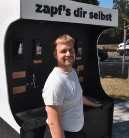 Ein lächelnder Ivan Malik steht im Freien vor einer tapeer Getränke-Zapfstation mit dem Schriftzug „zapf's dir selbst“.