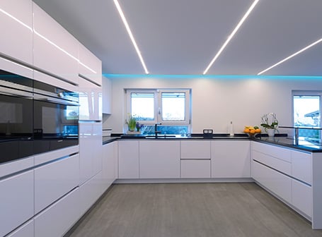 Eine moderne, weiße Hochglanzküche mit schwarzen Arbeitsplatten, die durch helle LED-Stripes beleuchtet wird, welche in die Decke eingelassen sind und für eine futuristische, akzentuierte Beleuchtung sorgen.