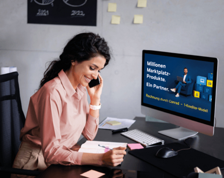 Eine lächelnde Frau sitzt am Schreibtisch, telefoniert mit einem Smartphone und macht sich Notizen, während auf dem Monitor vor ihr ein Angebot von Conrad angezeigt wird.