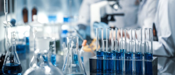 Abb. Chemisches Labor mit Reagenzgläsern, blauer Flüssigkeit und unscharfen Personen im Hintergrund