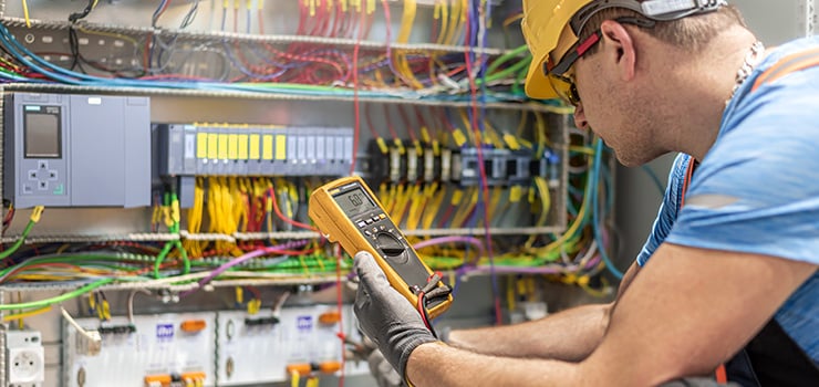 Elektriker misst mit einem Multimeter an einem Schaltschrank mit zahlreichen farbigen Kabeln und Steuerkomponenten.