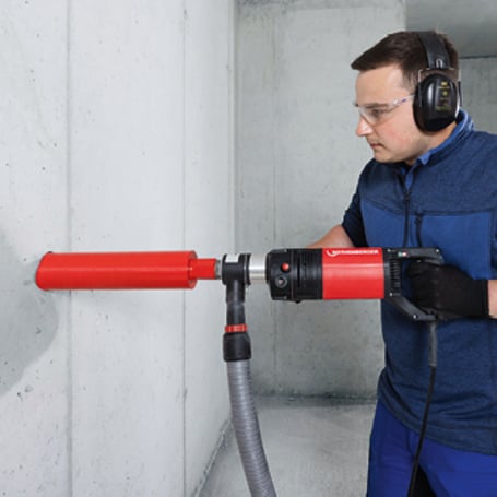Ein Handwerker mit Gehörschutz und Schutzbrille bohrt mit einer roten Kernbohrmaschine mit Staubabsaugung ein horizontales Loch in eine Betonwand.
