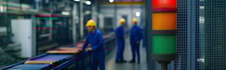 Drei Bauarbeiter in blauer Kleidung und gelben Helmen in einer Fabrik. Rechts eine Ampel mit rotem, gelbem und grünem Licht.