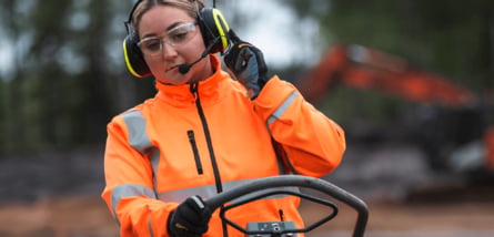Eine Arbeiterin in oranger Warnjacke mit Gehörschutz-Headset und Schutzbrille bedient eine Baumaschine auf einer Baustelle mit einem Bagger im Hintergrund.