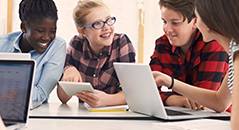 Ein Teaserbild zeigt vier lächelnde junge Menschen, die an einem Tisch sitzen und gemeinsam einen Laptop und ein Tablet benutzen, was eine kollaborative und interaktive Lernsituation darstellt