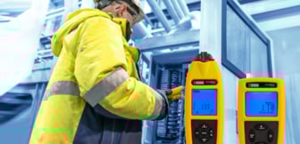  Ein Techniker in gelber Arbeitskleidung und mit einem Schutzhelm bedient ein elektrisches Messgerät an einem Schaltschrank, während zwei weitere gelbe Messgeräte mit blauen Displays im Vordergrund sichtbar sind.