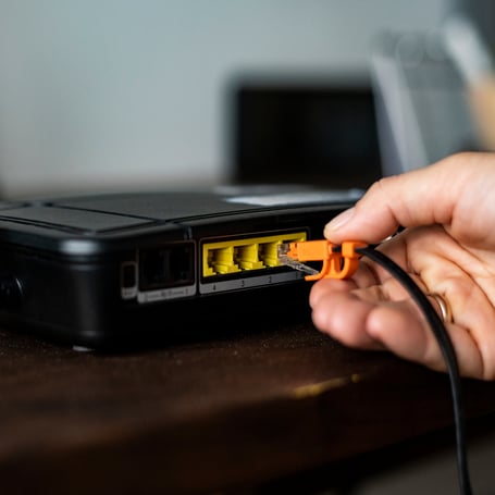 Eine Hand steckt ein schwarzes Netzwerkkabel mit orangefarbenem Stecker in den gelben LAN-Anschluss eines schwarzen Routers.