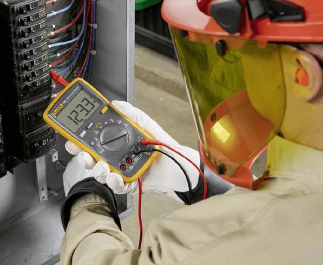 Ein Elektriker mit Schutzkleidung und Visier misst mit einem gelben Digital-Multimeter die Spannung an einer offenen Schalttafel mit zahlreichen Sicherungen.