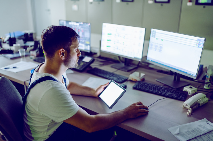 Ein Techniker in Arbeitskleidung sitzt in einem Kontrollraum vor mehreren Monitoren mit komplexen Systemanzeigen. In der einen Hand hält er ein Tablet, mit der anderen bedient er eine Computermaus.