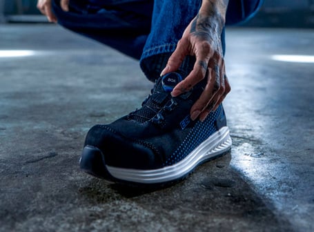 Eine Person in hockender Position bedient mit tätowierter Hand den blauen BOA-Drehverschluss eines modernen, schwarz-blauen ELTEN Sicherheitsschuhs auf einem grauen Betonboden.