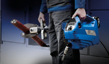 Eine Person in Arbeitskleidung trägt eine tragbare blaue Akku-Hydraulikpumpe, die über einen Schlauch mit einem hydraulischen Schneidkopf an einem roten Kabel verbunden ist.