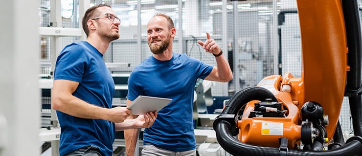 Abb. Zwei Männer in einem industriellen Umfeld diskutieren vor einem Industrieroboter, einer hält ein Tablet