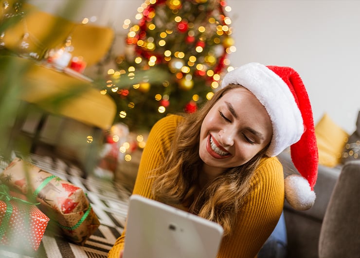 EIne Frau liegt im Wohnzimmer auf einem Teppich mit einem Tablet in der Hand und einer Weihnachtsmannmütze auf dem Kopf. Im Hintergrund ist es weihnachtlich dekoriert mit einem leuchtenden Weihnachtsbaum und eingepackten Geschenken.