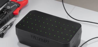 Ein schwarzes Voltcraft Rundzellen-Ladegerät mit zahlreichen leuchtenden grünen LEDs auf der Oberseite steht neben Werkzeugen wie einer Stromzange und einem Schraubendreher auf einer hellen Arbeitsfläche.