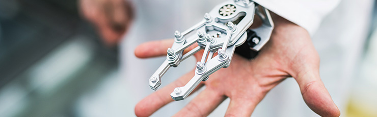 Nahaufnahme einer menschlichen Hand, an der eine mechanische, silberne Roboter-Exoskelett-Hand mit Gelenken und Schrauben befestigt ist, die wie eine technische Greifhilfe über den Fingern liegt.