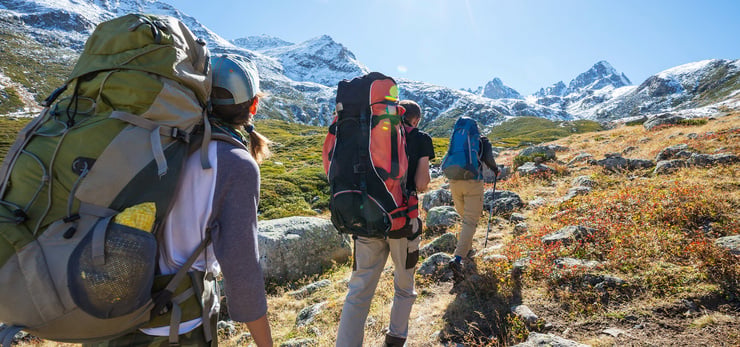 Wandergruppe mit grossen Rucksäcken unterwegs in einer alpinen Berglandschaft mit schneebedeckten Gipfeln.