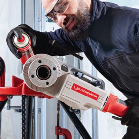 Ein Handwerker mit Schutzbrille sprüht Schneidöl auf eine elektrische Gewindeschneidmaschine, die ein Gewinde auf ein Rohr schneidet.