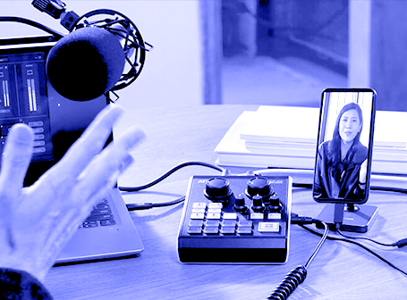 Eine monochrome, blau eingefärbte Nahaufnahme eines Streaming- oder Podcast-Setups auf einem Tisch, bei dem neben einer gestikulierenden Hand, einem Mikrofon und einem Laptop ein kompaktes Audio-Mischpult sowie ein Smartphone im Ständer mit einer Person auf dem Display zu sehen sind.